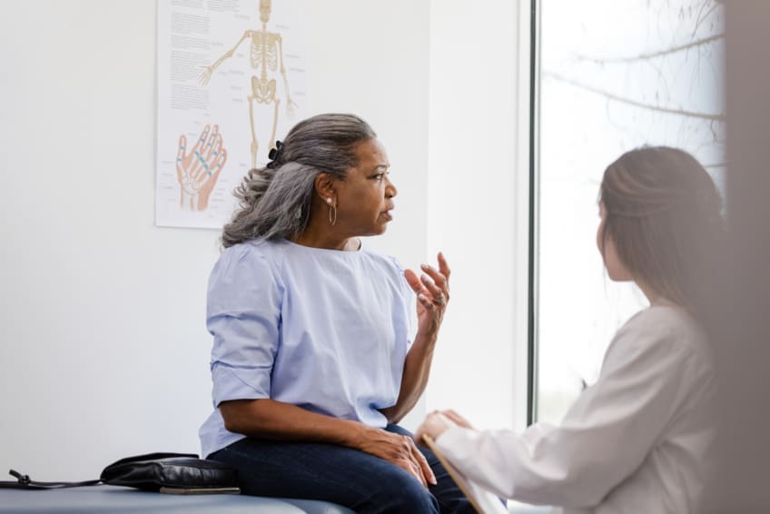 A woman, appearing worried, is seated on an exam table while talking with her doctor.